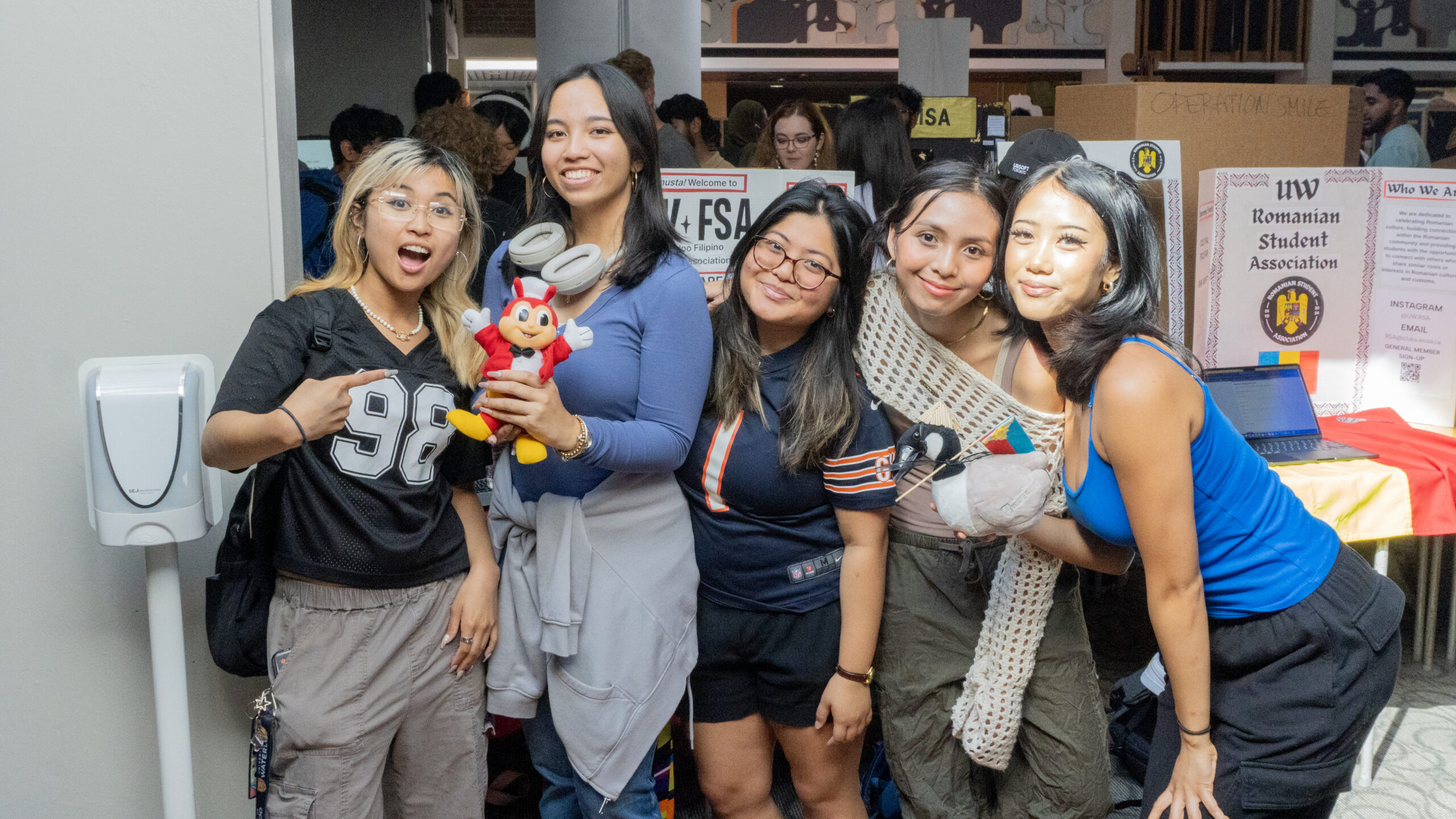 Five young women smile and pose together indoors at an event, standing in front of display tables. Showcasing free UW tools to boost your semester, they hold a red plush toy and a stuffed animal. A crowd and informational posters are visible behind them.