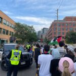 A group of people march down a city street holding Palestinian flags. Police officers in reflective vests and a police vehicle are present. Brick buildings and trees line the road under a partly cloudy sky.