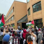 A large crowd gathers outside a brick building holding Palestinian flags and protest signs, with many people wearing keffiyehs. Some speakers address the crowd from the building entrance. The atmosphere appears peaceful but passionate.