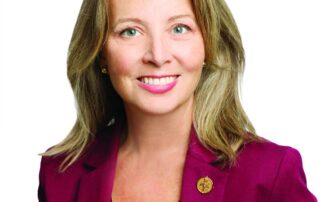 A woman with shoulder-length blonde hair, wearing a burgundy blazer over a black top, smiles at the camera against a white background—capturing Marit Stiles during the All in for Ontario Tour.