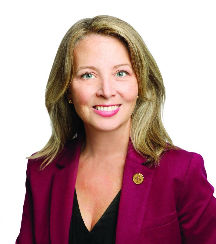 A woman with shoulder-length blonde hair, wearing a burgundy blazer over a black top, smiles at the camera against a white background—capturing Marit Stiles during the All in for Ontario Tour.