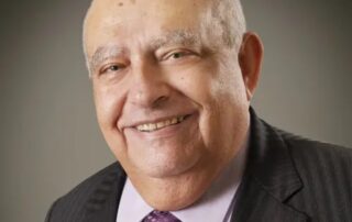 An older man with a bald head smiles at the camera. Wearing a dark suit, light purple shirt, and purple patterned tie, the distinguished dean of engineering is set against a neutral background.