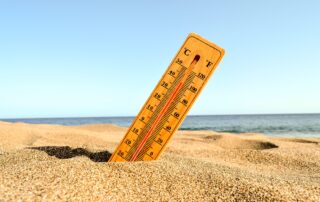 A wooden thermometer stuck in sandy beach shows a high temperature under a clear blue sky, with the ocean visible in the background, highlighting the need for heat warnings and weather alerts during hot summer days.