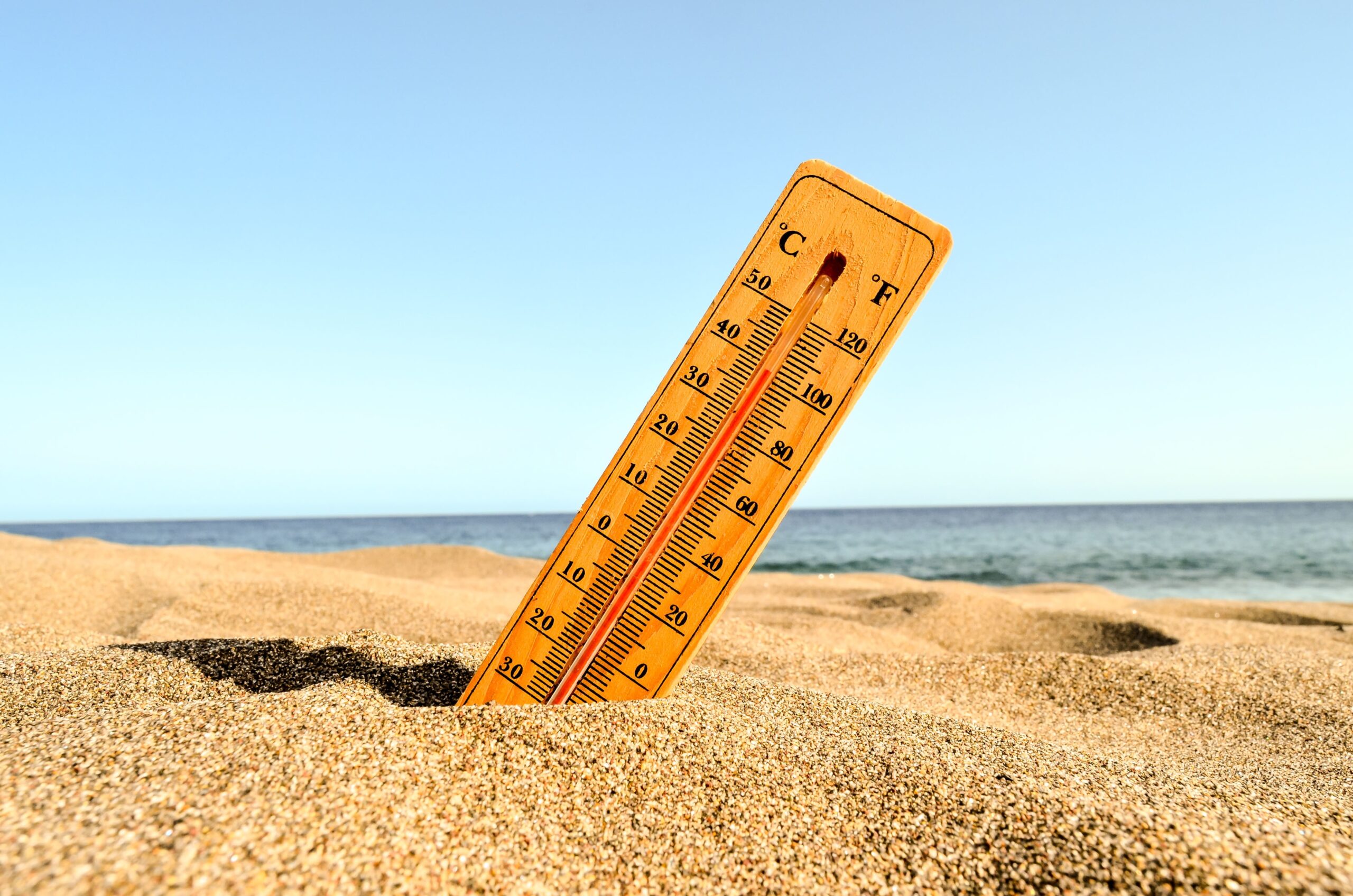 A wooden thermometer stuck in sandy beach shows a high temperature under a clear blue sky, with the ocean visible in the background, highlighting the need for heat warnings and weather alerts during hot summer days.
