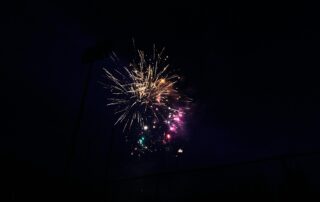 Colorful fireworks charges burst in the night sky over Waterloo region, illuminating the darkness with bright yellow, pink, purple, and green sparks. A faint outline of a fence and stadium lights is visible below.