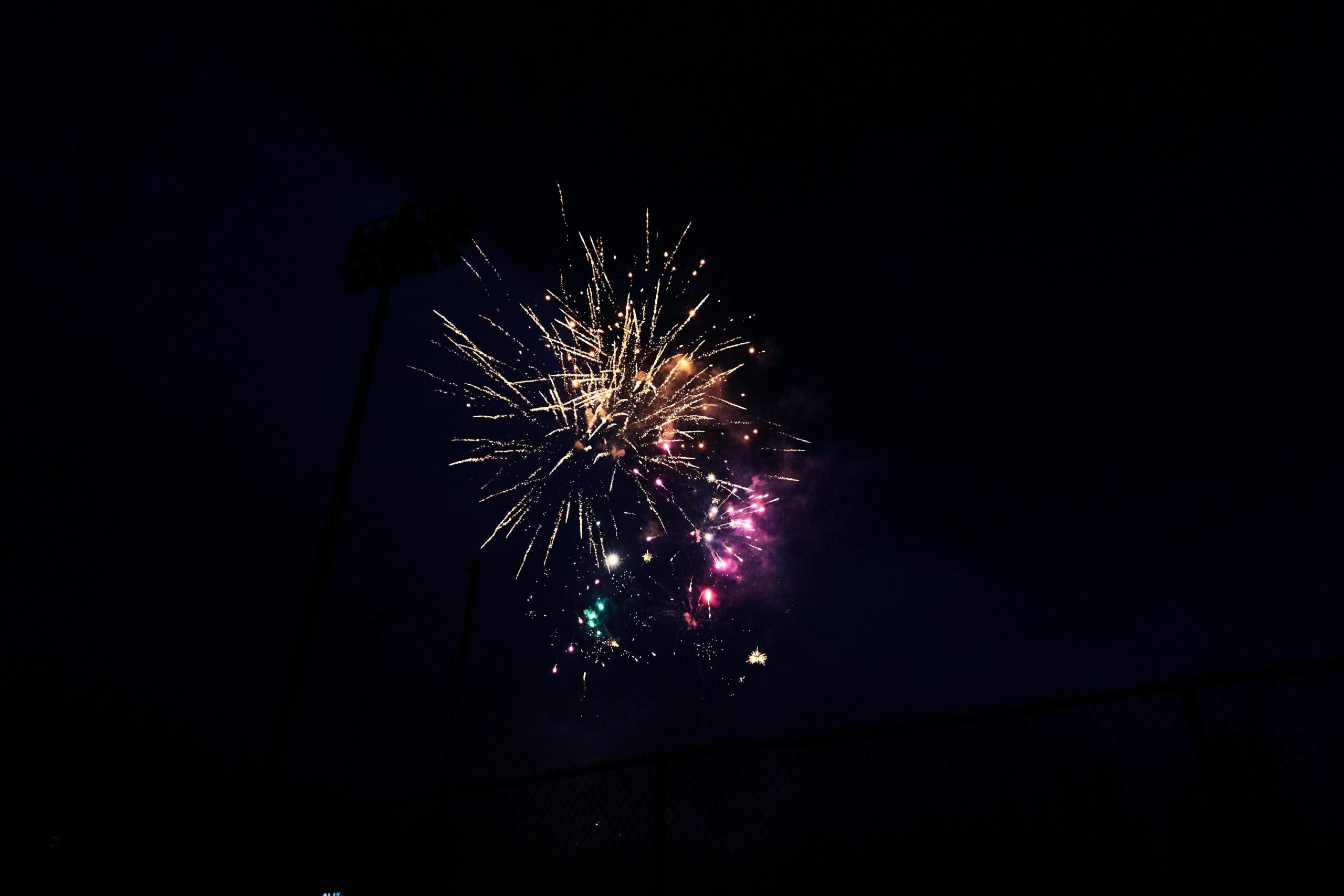 Colorful fireworks charges burst in the night sky over Waterloo region, illuminating the darkness with bright yellow, pink, purple, and green sparks. A faint outline of a fence and stadium lights is visible below.