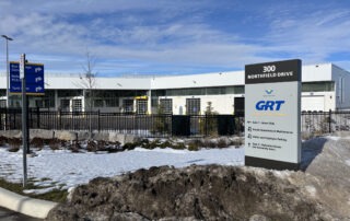 A modern building with a “300 Northfield Drive” sign for GRT, Grand River Transit, stands behind a snowy, slushy sidewalk in Waterloo. Directional signs help with navigating to nearby cities under a partly cloudy sky.