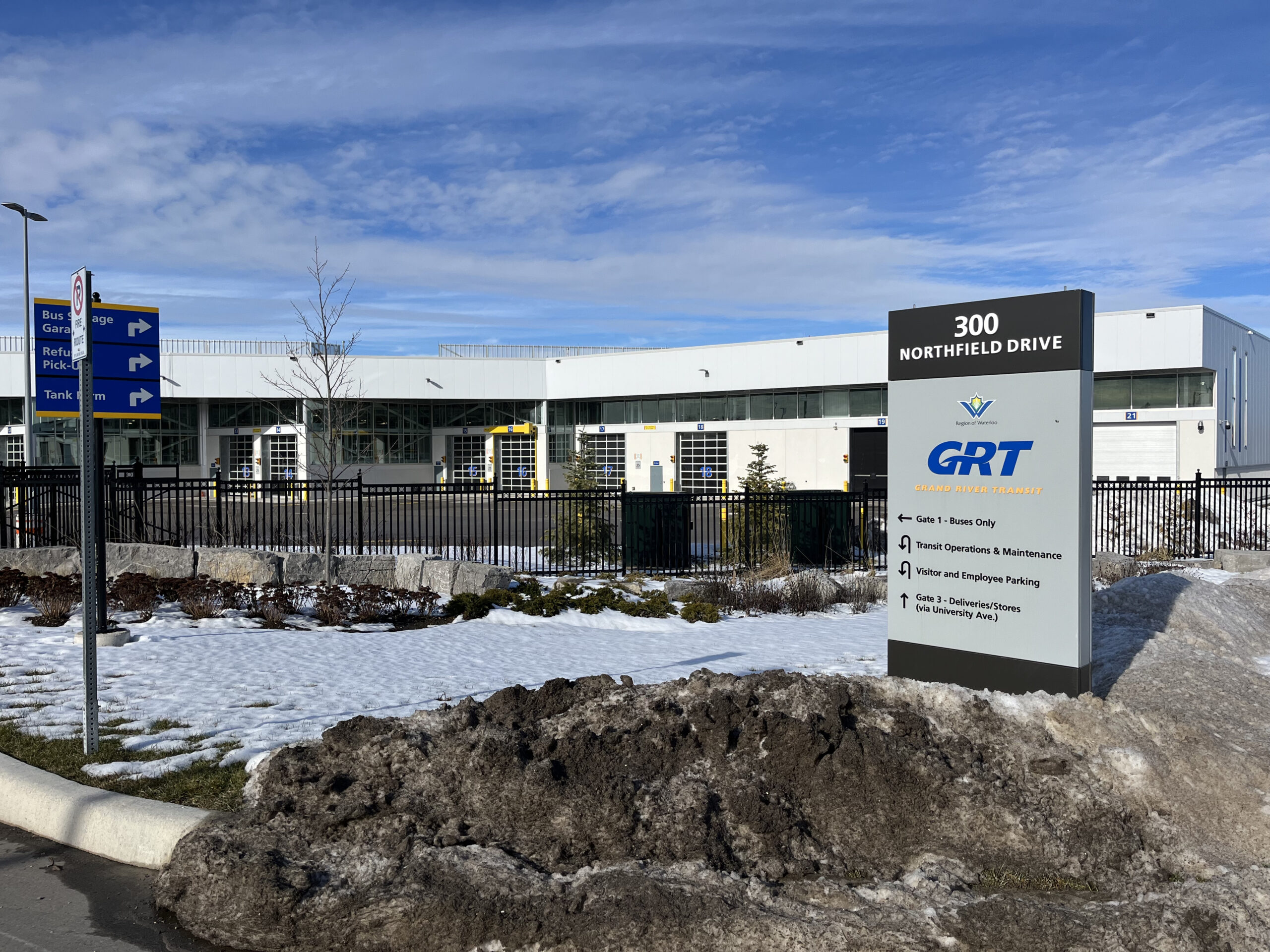 A modern building with a “300 Northfield Drive” sign for GRT, Grand River Transit, stands behind a snowy, slushy sidewalk in Waterloo. Directional signs help with navigating to nearby cities under a partly cloudy sky.