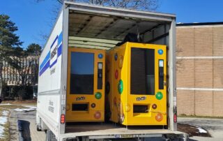 Two large yellow M&M's vending machines featuring colorful candy images are inside the back of an open truck parked outdoors near a UW building, with some snow on the ground—raising questions about vending machine privacy.