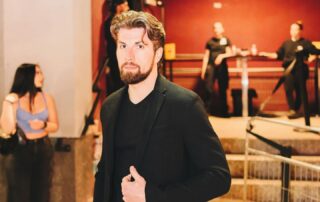 A man with brown hair and a beard, possibly Andrei Nicola, known as the King of Waterloo, stands near a staircase at an indoor event. Other people are visible in the background. Yellow text reads "#Rose4Kha" in the bottom right corner.