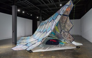 A large, tent-like installation made of colorful, translucent panels is displayed in a gallery. The structure drapes across the floor, with cushions placed underneath, inviting visitors to relax or even take naps in the cozy, shelter-like space.