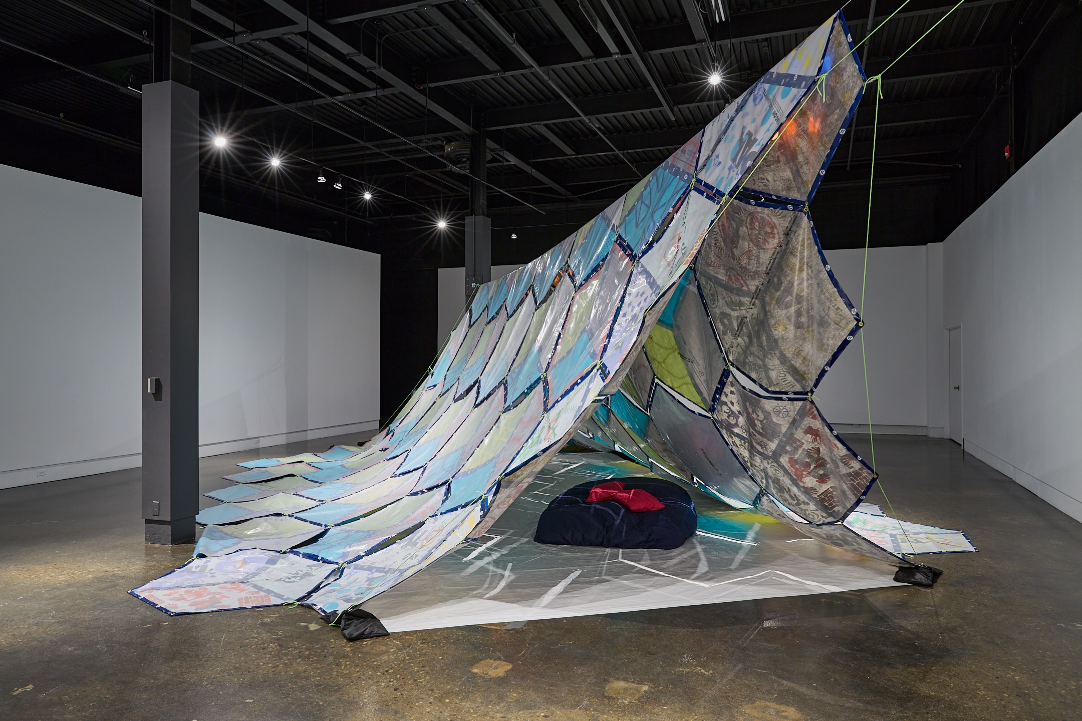 A large, tent-like installation made of colorful, translucent panels is displayed in a gallery. The structure drapes across the floor, with cushions placed underneath, inviting visitors to relax or even take naps in the cozy, shelter-like space.