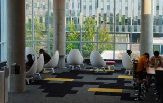 A modern lounge area—one of the best study spots—features people in white, egg-shaped chairs and at a long table. Large windows reveal trees and a geometric office building, while the floor boasts black and yellow geometric patterns.