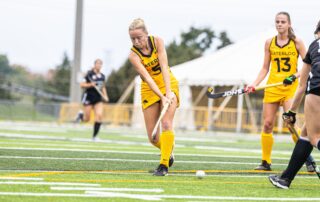 A female field hockey player in a yellow "Waterloo" uniform, showcasing Meagan Hobson’s legacy of leadership, prepares to hit the ball during a game, with teammates and opponents visible in the background on the turf field.