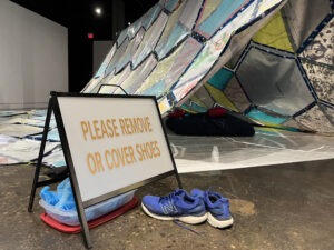A sign reading "PLEASE REMOVE OR COVER SHOES" stands next to a pair of blue sneakers and shoe covers, inviting visitors to keep the cozy, colorful tent-like structure indoors clean—a perfect spot for quiet naps.