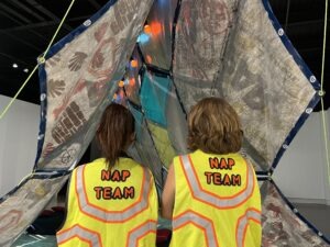 Two people with light brown hair wear yellow vests labeled "NAP TEAM" as they face a large, tent-like structure covered in colorful handprints and designs, highlighting the art of naps inside an art gallery.