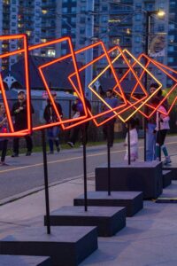 A row of glowing orange neon square frames stands outdoors at dusk during Lumen 2025, with people catching a glimpse of the art installation. Tall buildings and a chain-link fence are visible in the sparkling night background.