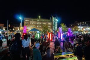 A large crowd gathers at an outdoor plaza at night for Lumen 2025, illuminated by colorful, glowing light pillars and sculptures. People mingle as surrounding buildings light up the sparkling night against the dark sky.