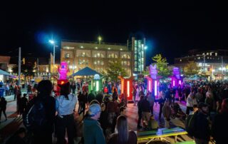 A large crowd gathers at an outdoor plaza at night for Lumen 2025, illuminated by colorful, glowing light pillars and sculptures. People mingle as surrounding buildings light up the sparkling night against the dark sky.
