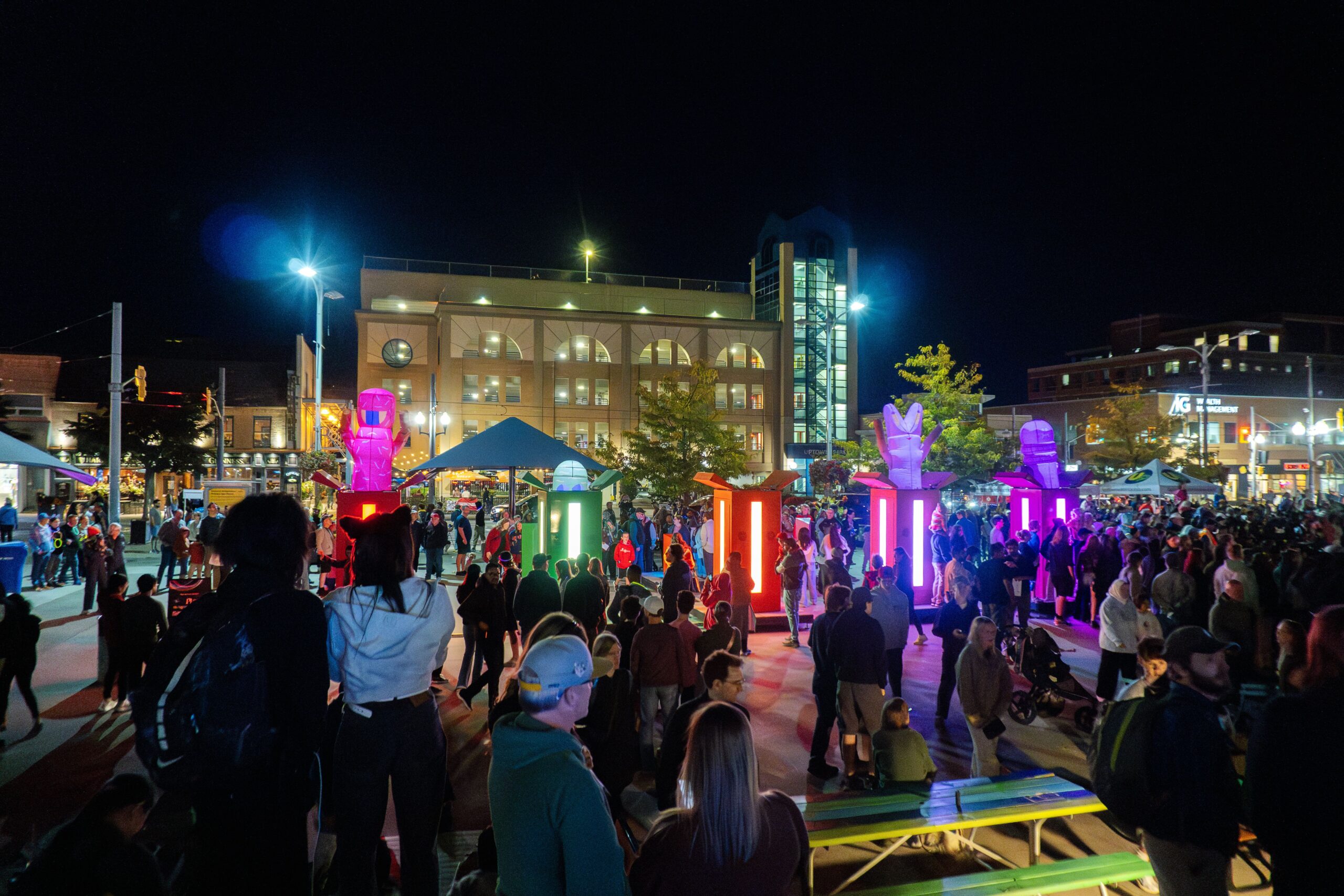 A large crowd gathers at an outdoor plaza at night for Lumen 2025, illuminated by colorful, glowing light pillars and sculptures. People mingle as surrounding buildings light up the sparkling night against the dark sky.