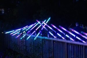 People stand on a bridge at night, illuminated by sparkling neon light bars arranged in vibrant, intersecting patterns for Lumen 2025, casting blue, pink, and purple hues across the dark surroundings.