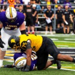 A Golden Hawks football player in a yellow and black uniform tackles a Warriors player in purple, pinning him to the turf as another Warrior watches during the intense Battle of Waterloo. Spectators and team members are visible in the background.