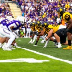 Two American football teams, the Warriors in white and purple and the Golden Hawks in black and yellow, face off on the line of scrimmage during a game, with a crowd watching their own Battle of Waterloo unfold in the background.