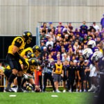 Two American football teams, the Warriors in white and purple and the Golden Hawks in yellow and black, line up on the field as the play is about to begin, with a crowd of spectators watching this modern Battle of Waterloo.