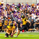 A football player in a yellow and black Golden Hawks uniform kicks a football held by a teammate during a game, with a crowd of spectators in the stands behind them.