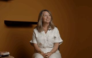 A woman with shoulder-length light hair, wearing a white outfit, sits smiling and looking up. In the orange-brown room with dark shelves, another person’s hand holding a notebook is visible—perhaps discussing government fair share or Marit Stiles.