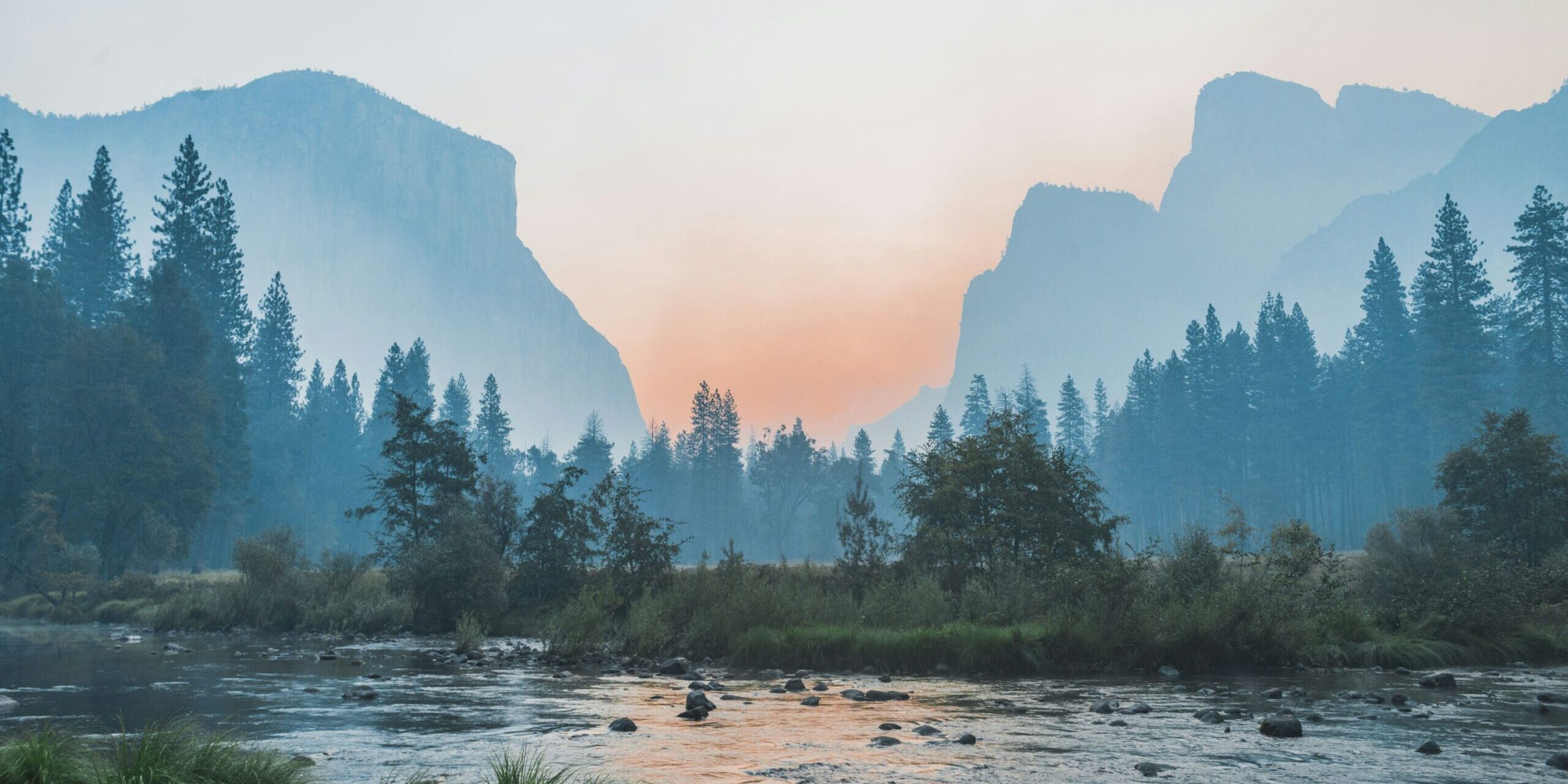 A serene river flows through a lush valley with tall pine trees, surrounded by towering mountain cliffs under a hazy, pastel-colored sky—capturing the tranquility of sunrise or sunset.