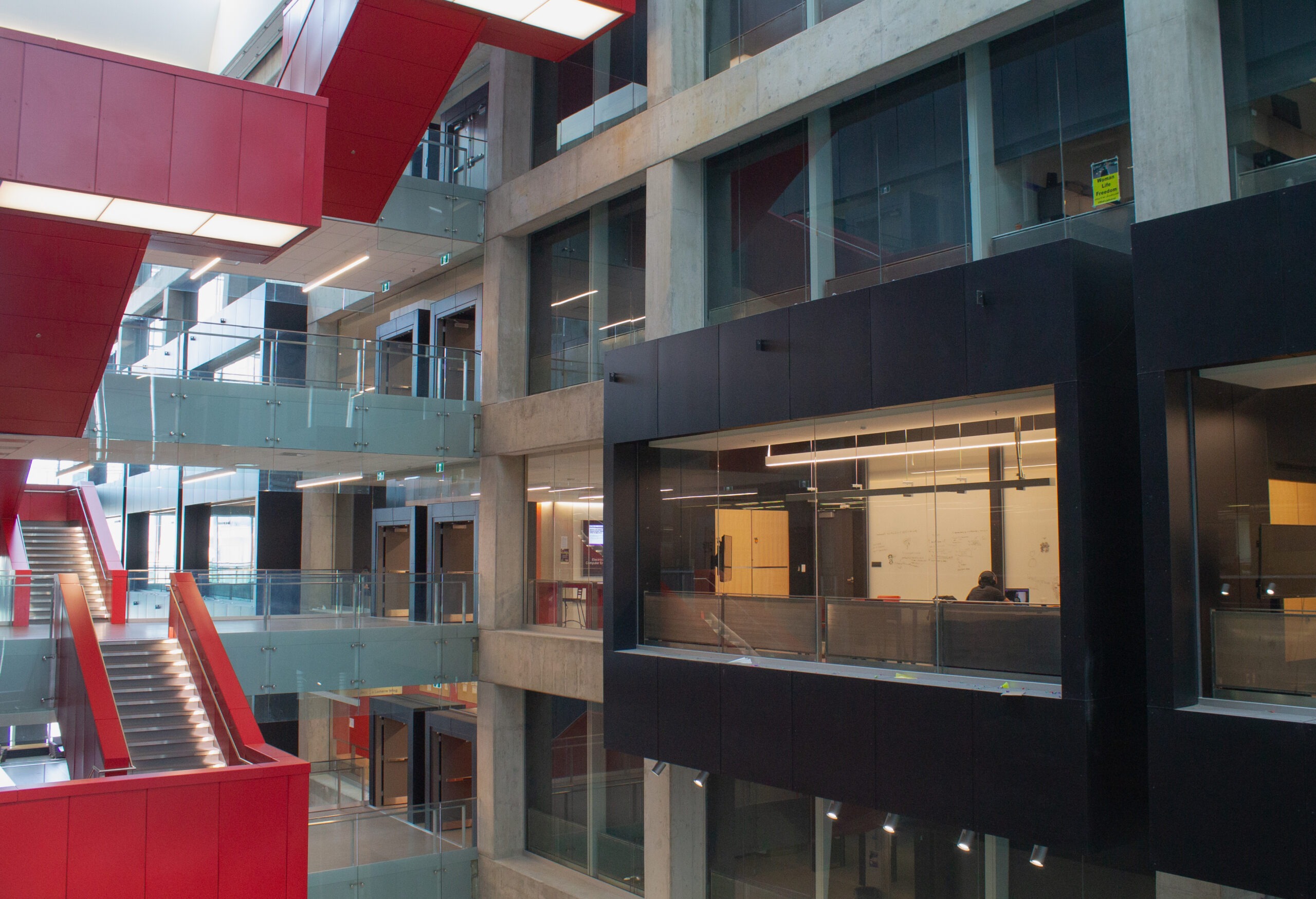 A modern building interior on the UW campus with large glass windows, concrete walls, red staircases, and suspended walkways connecting multiple floors—an open, spacious design ideal for a student advocacy group’s activities.