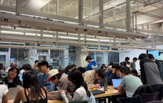 A large group of people sit and stand around tables in a modern, industrial-style room, many working on laptops and talking in groups during an Electrium Mobility orientation focused on a hands-on project under bright ceiling lights and exposed pipes.