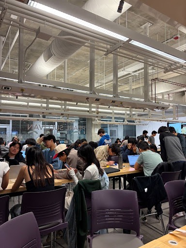 A large group of people sit and stand around tables in a modern, industrial-style room, many working on laptops and talking in groups during an Electrium Mobility orientation focused on a hands-on project under bright ceiling lights and exposed pipes.