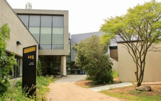 A modern building labeled "HS Health Services" with a large glass entrance, surrounded by greenery, trees, and a winding gravel path.
