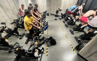 A group of people participate in a spin class, riding stationary bikes in a concrete-walled room. They are focused on their workout, perhaps training for the upcoming Ring Road Rumble or gearing up for their next UW triathlon. Gym bags are visible on the floor.