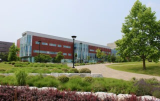 A modern, three-story brick and glass building stands behind landscaped gardens, a stone border, and a curving paved walkway, with trees and shrubs under a clear sky.