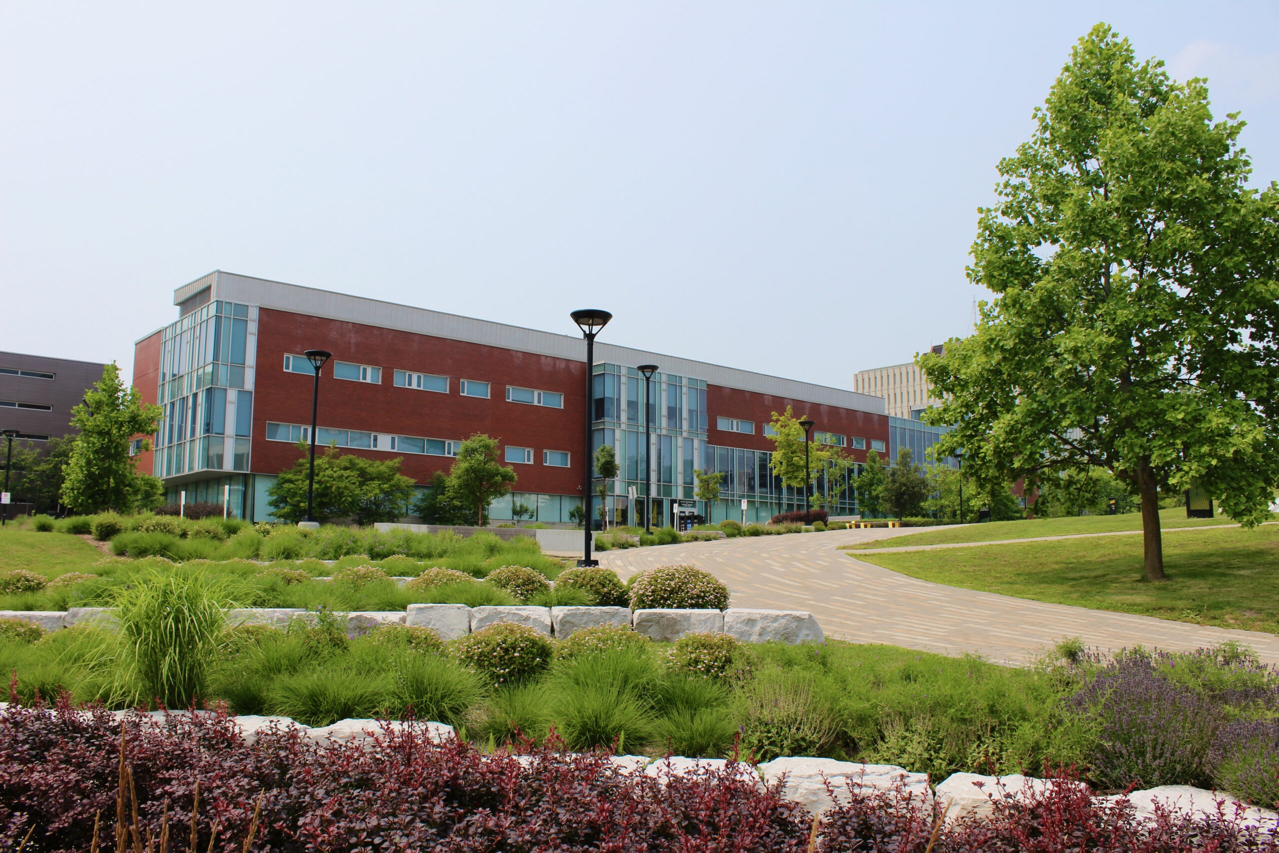 A modern, three-story brick and glass building stands behind landscaped gardens, a stone border, and a curving paved walkway, with trees and shrubs under a clear sky.