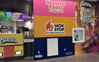 A vibrant food court wall in SLC features colorful signage for "Smarty Pants Ice Cream & More," "Wok Stop Fast Fresh Wok'd To Go," and "Chaska Indian Street Food Obsession." A counter and digital menu are partially visible on the left.