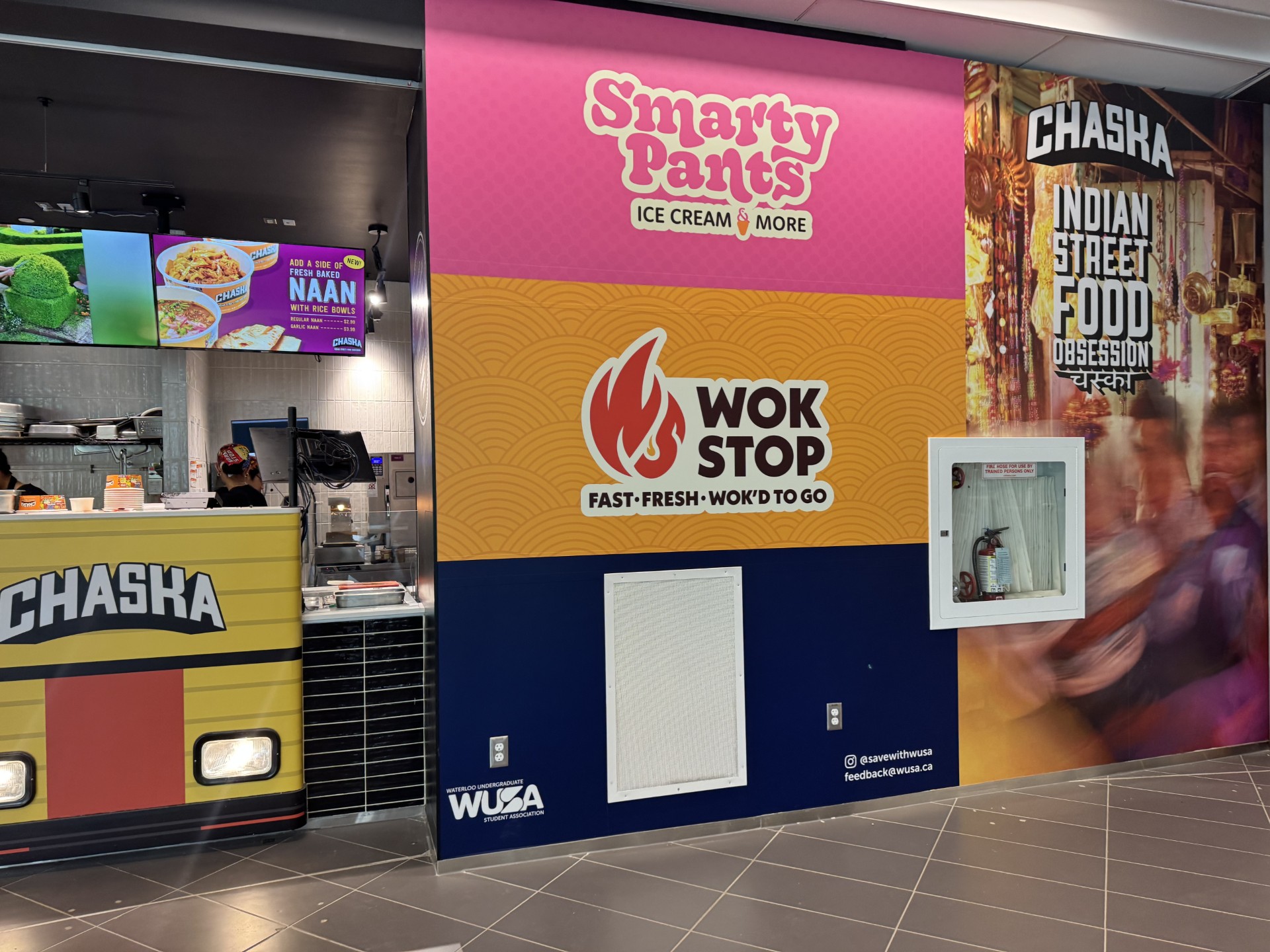 A vibrant food court wall in SLC features colorful signage for "Smarty Pants Ice Cream & More," "Wok Stop Fast Fresh Wok'd To Go," and "Chaska Indian Street Food Obsession." A counter and digital menu are partially visible on the left.