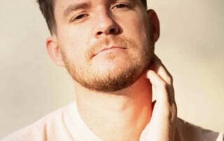 A young man with short, curly brown hair and a beard, styled in the manner of digital art, looks into the camera while touching his neck. He is wearing a light-colored shirt and is lit by soft, natural light against a pale background.