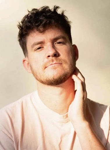 A young man with short, curly brown hair and a beard, styled in the manner of digital art, looks into the camera while touching his neck. He is wearing a light-colored shirt and is lit by soft, natural light against a pale background.