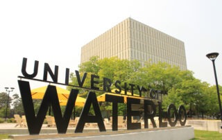 Large black "University of Waterloo" sign in front of yellow umbrellas and patio tables, with a tall, beige rectangular building and green trees in the background—perfect for checking library hours or relaxing between study sessions.