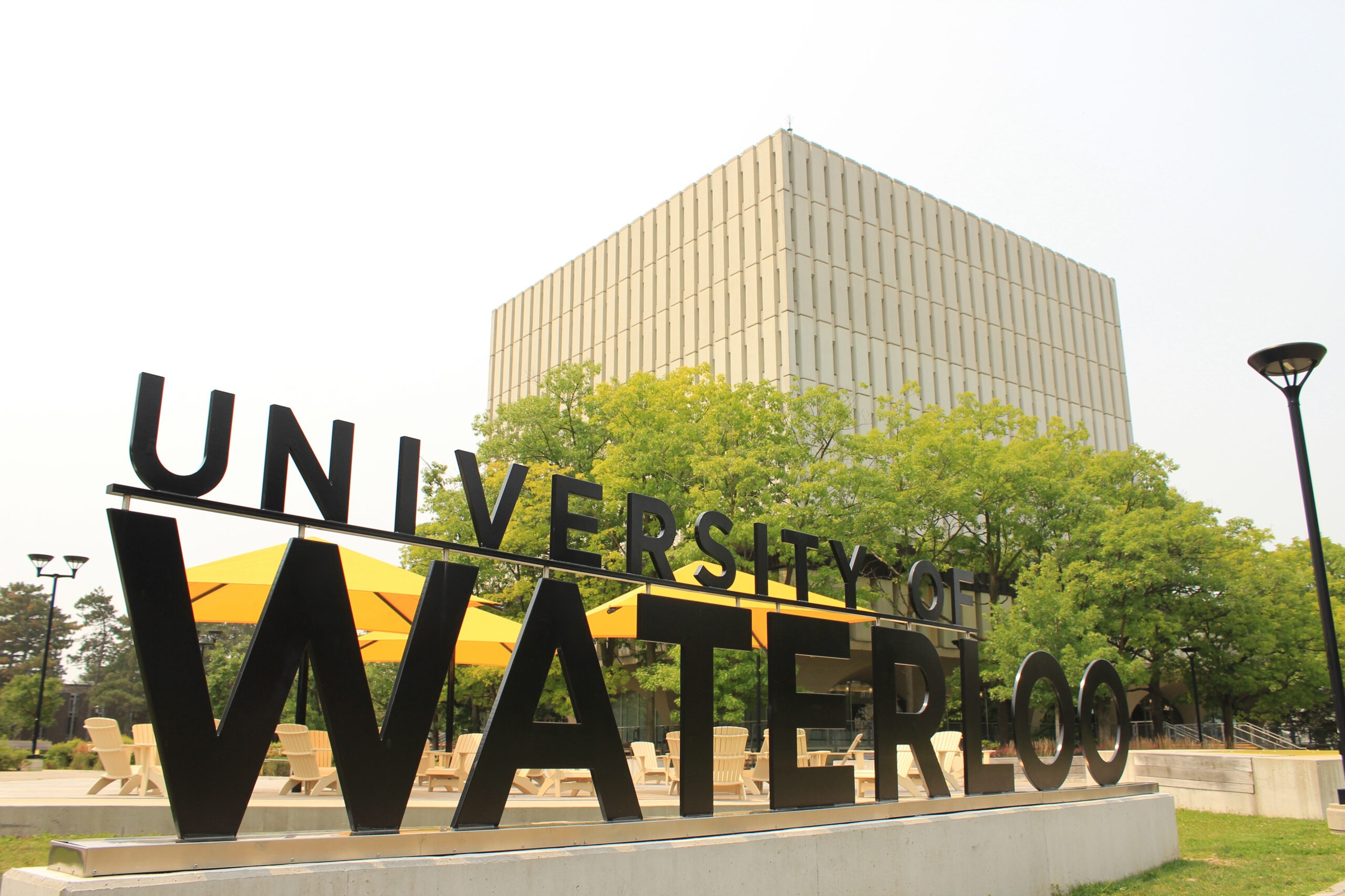 Large black "University of Waterloo" sign in front of yellow umbrellas and patio tables, with a tall, beige rectangular building and green trees in the background—perfect for checking library hours or relaxing between study sessions.
