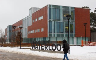 A person walks past a large "University of Waterloo" sign on the UW campus in front of a modern building with glass windows on a snowy, overcast day.
