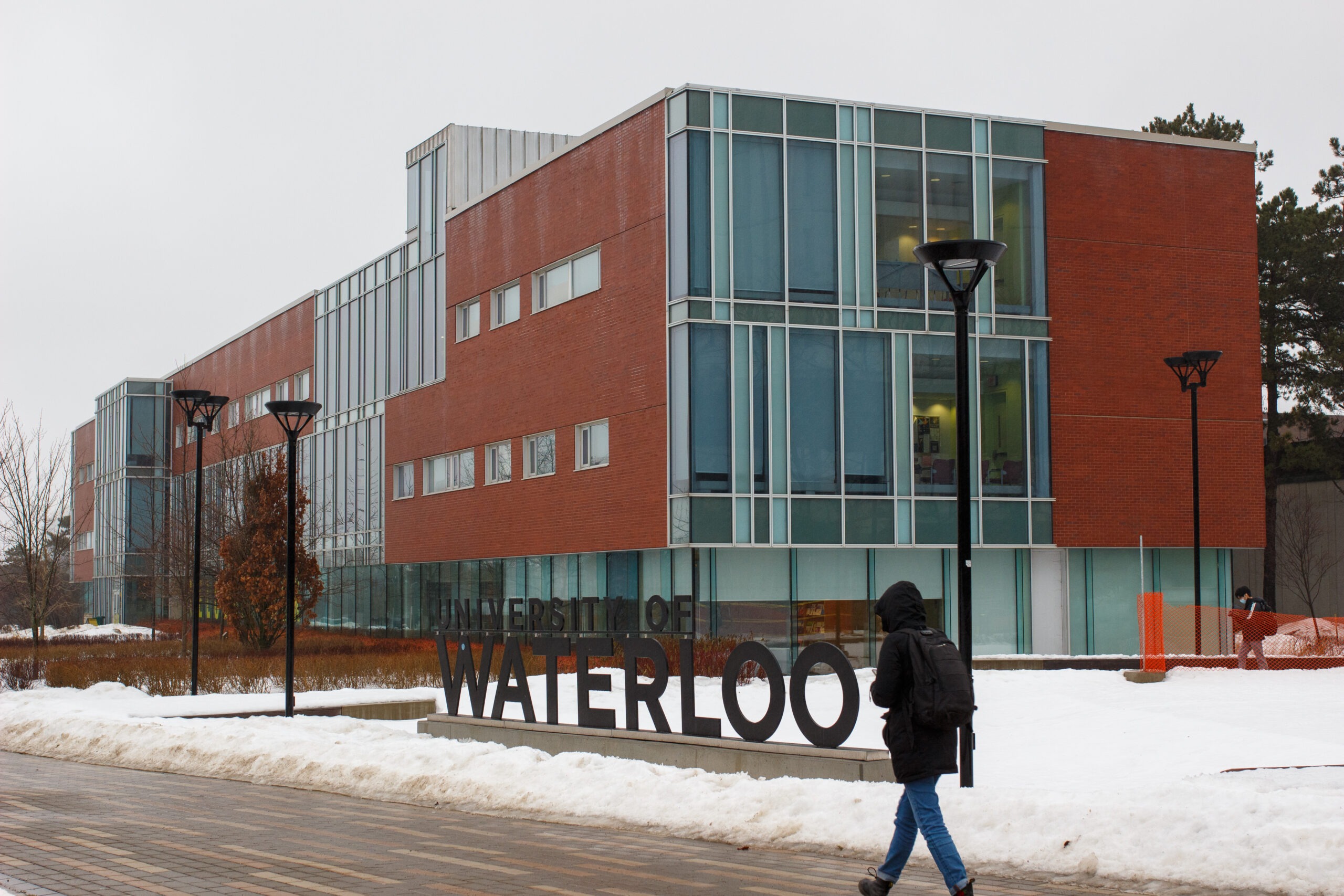 A person walks past a large "University of Waterloo" sign on the UW campus in front of a modern building with glass windows on a snowy, overcast day.