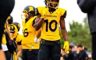 A football player in a yellow and black "Warriors" uniform with the number 10 walks on the field during the Homecoming game, surrounded by teammates. Water bottles are lined up on the sidelines and one person is clapping in the foreground.