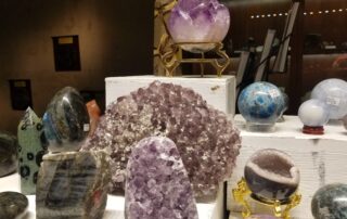 A display of polished and raw gemstones, including amethyst clusters, spherical stones, and geodes, arranged on white stands in a shop setting with soft lighting reminiscent of a Gem and Mineral Show.