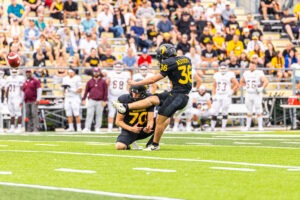 A football player in a black uniform, possibly one of the Athletes of the Week like Arthur Kowara, kicks a football held by a teammate during a game, as players and spectators watch from the background.