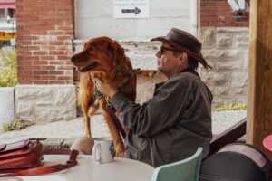 A man wearing sunglasses and a hat, possibly UW staff or a National Book Award-nominated author, sits at an outdoor table holding a brown dog on a leash. A mug, red bag, and other items rest on the table amid brick walls and a gravel path.